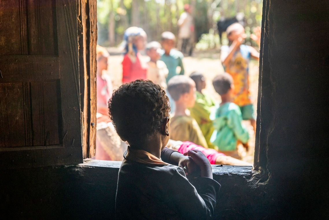 Ein kleiner Junge schaut aus dem Fenster eine Hütte nach draußen, wo Kinder spielen.