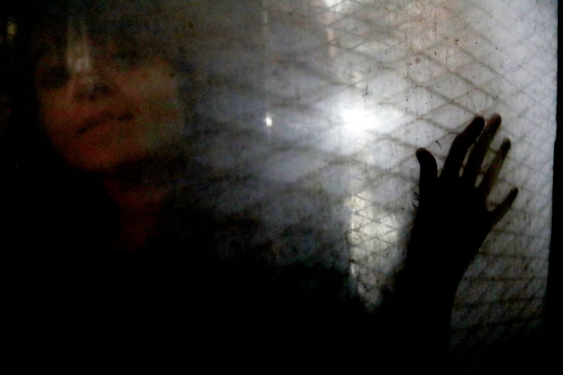 Gesicht einer Frau hinter einer schmutzigen Glasscheibe mit Rautenmuster. Sie legt ihre linke Hand auf die Scheibe.