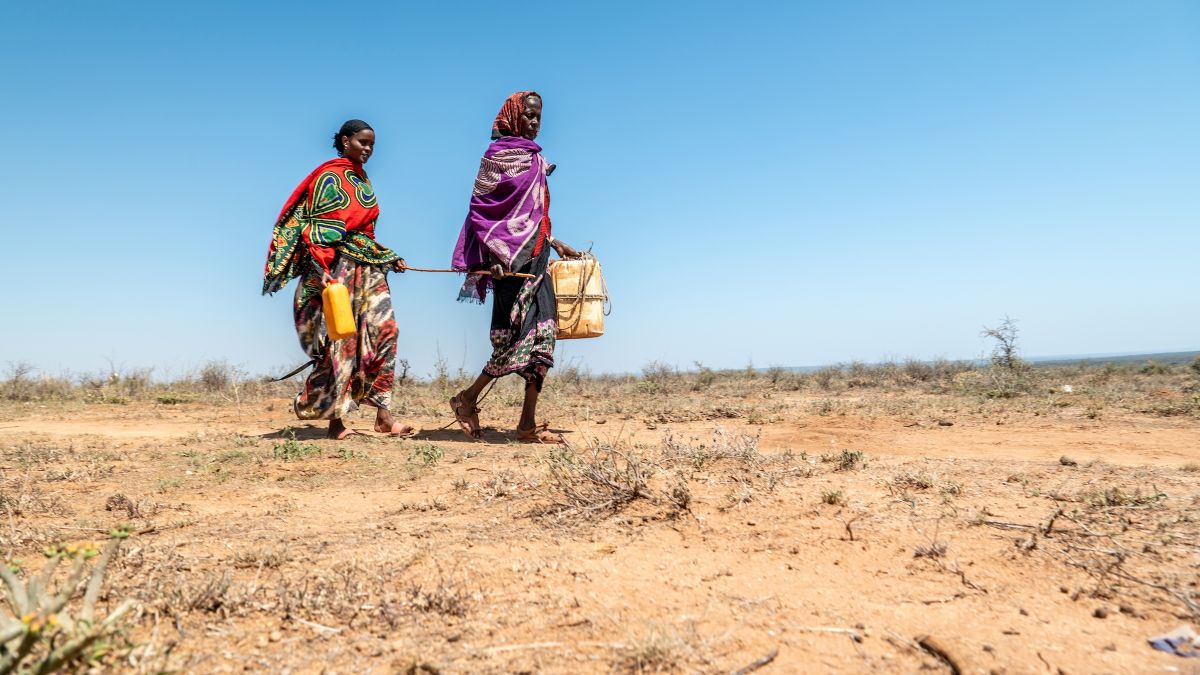 Eine ältere Frau mit buntem Kopftuch führt eine andere, jüngere Frau über gelben, trockenen, unbewachsenen Boden. Sie halten zum Führen jeweils ein Ende eines Stocks, der sie verbindet.