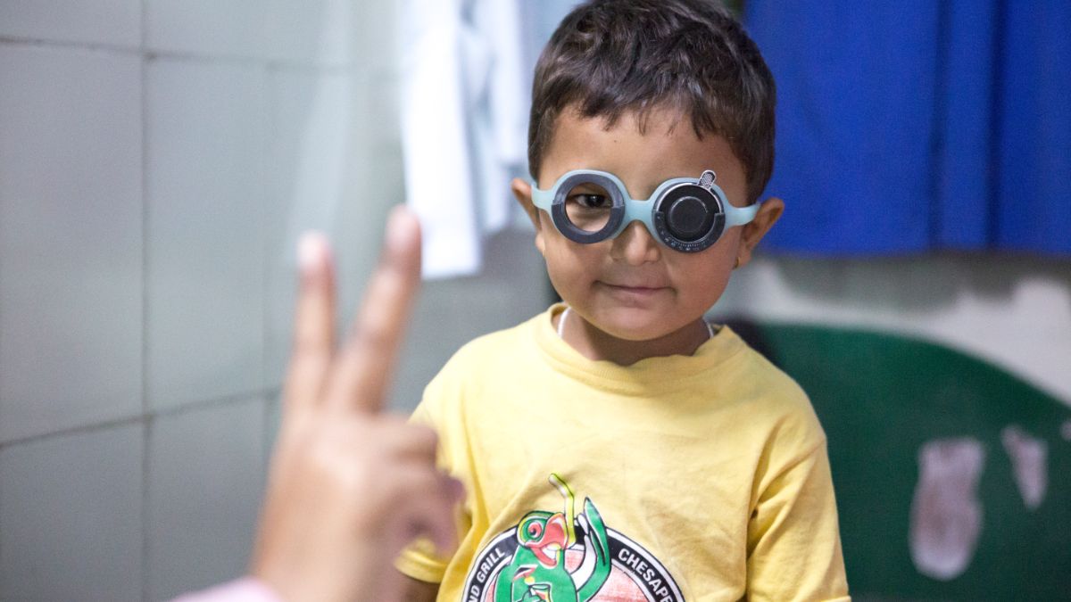 Kleiner Junge mit Brille auf den Augen. Vor ihm hält eine Person zwei Finger hoch.