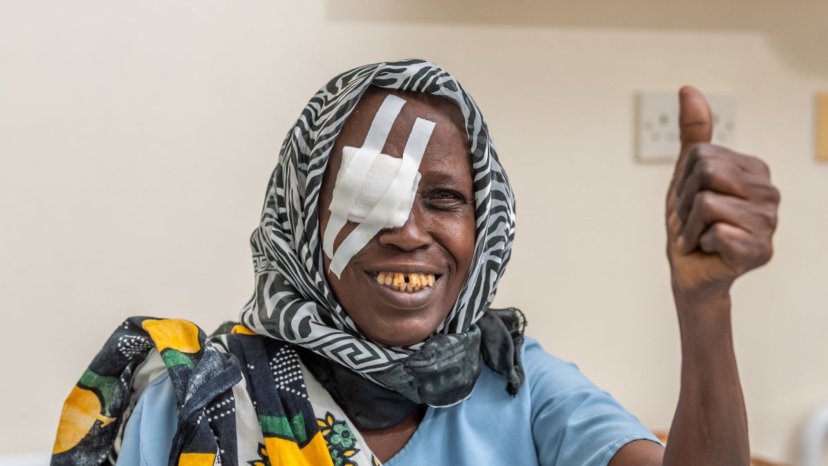 Frau mit Tuch über ihren Kopf trägt eine Augenklappe auf dem rechten Auge und hält ihren Daumen hoch.