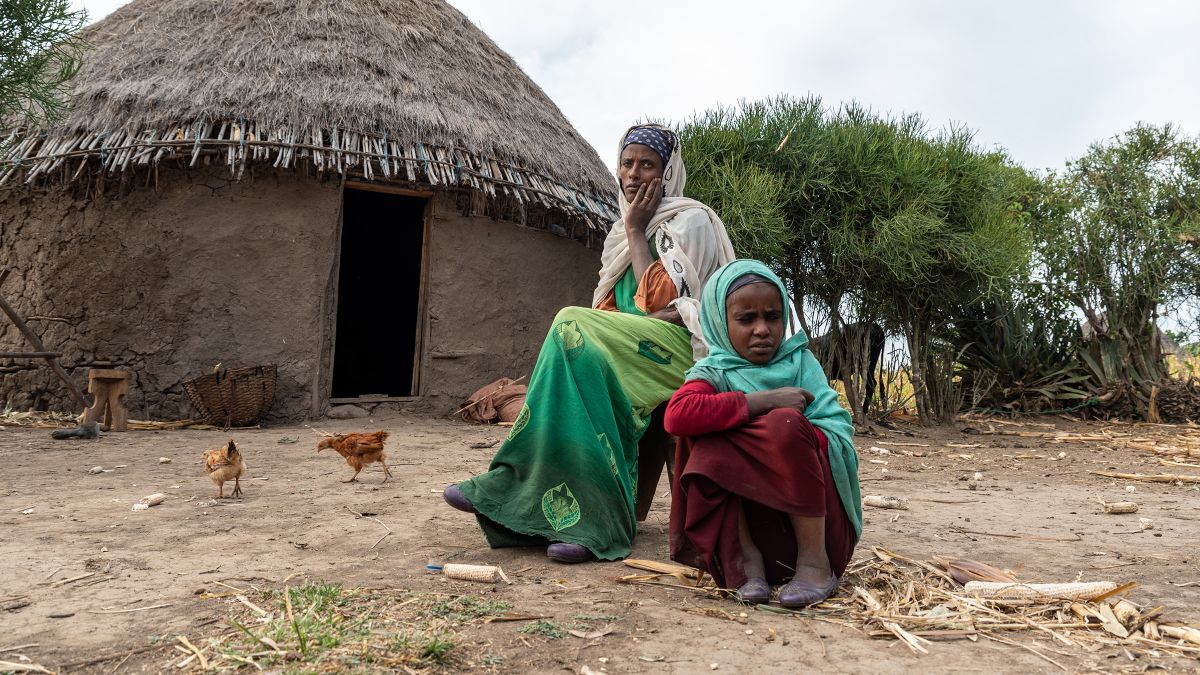 Eine Frau und ein kleines Mädchen sitzen auf dem Platz vor einer strohgedeckten, runden Lehmhütte. Einige Hühner laufen um sie herum.
