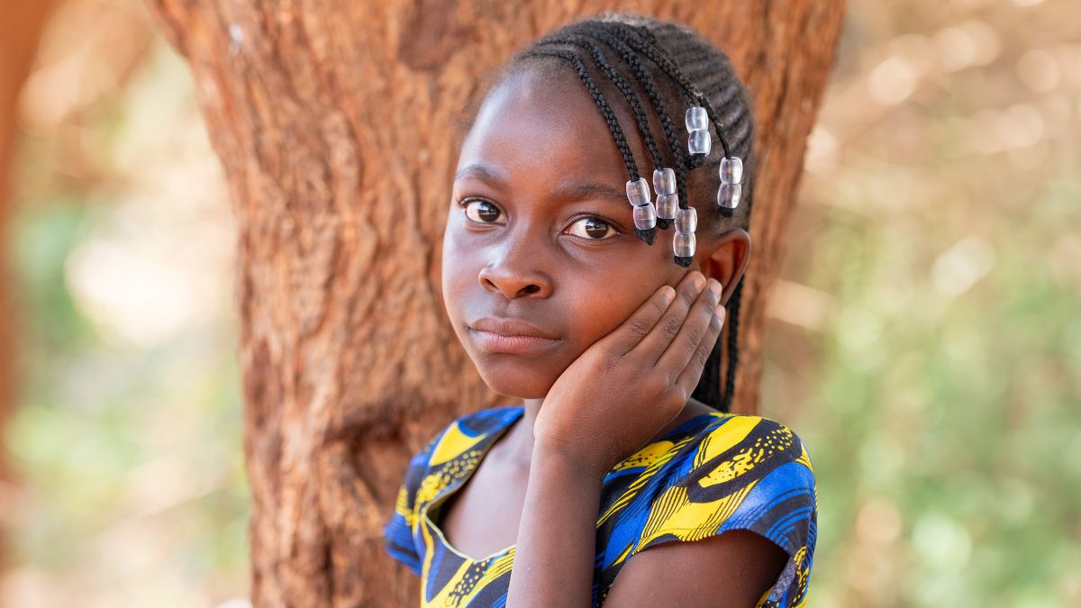Ein Mädchen mit kleinen Zöpfchen und Glasperlen im Haar steht vor einem Baum. Es legt die Hand ans linke Ohr.