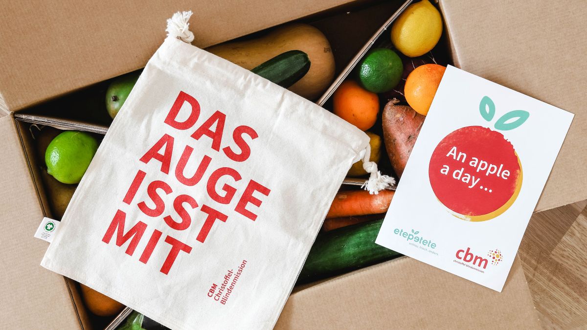 Offener Pappkarton voller Obst und Gemüse, darauf ein Stoffbeutel mit der Aufschrift &quot;Das Auge isst mit&quot;