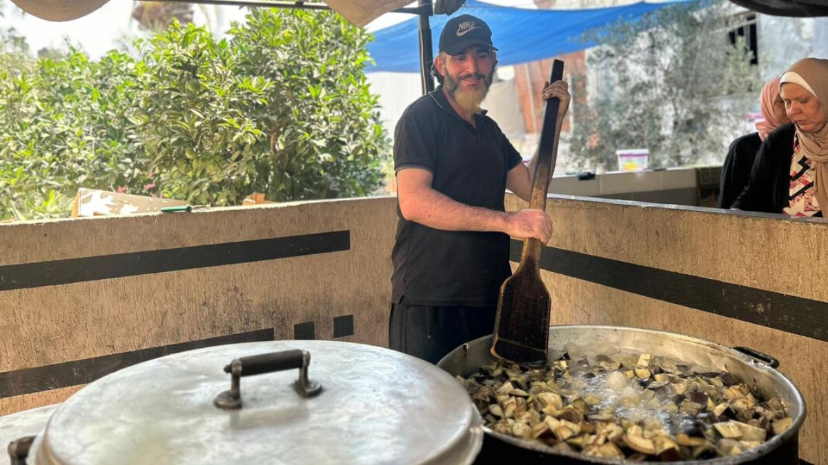 Ein Mann mit Schirmmütze und Bart rührt in einem großen Kochtopf mit Auberginenstücken.