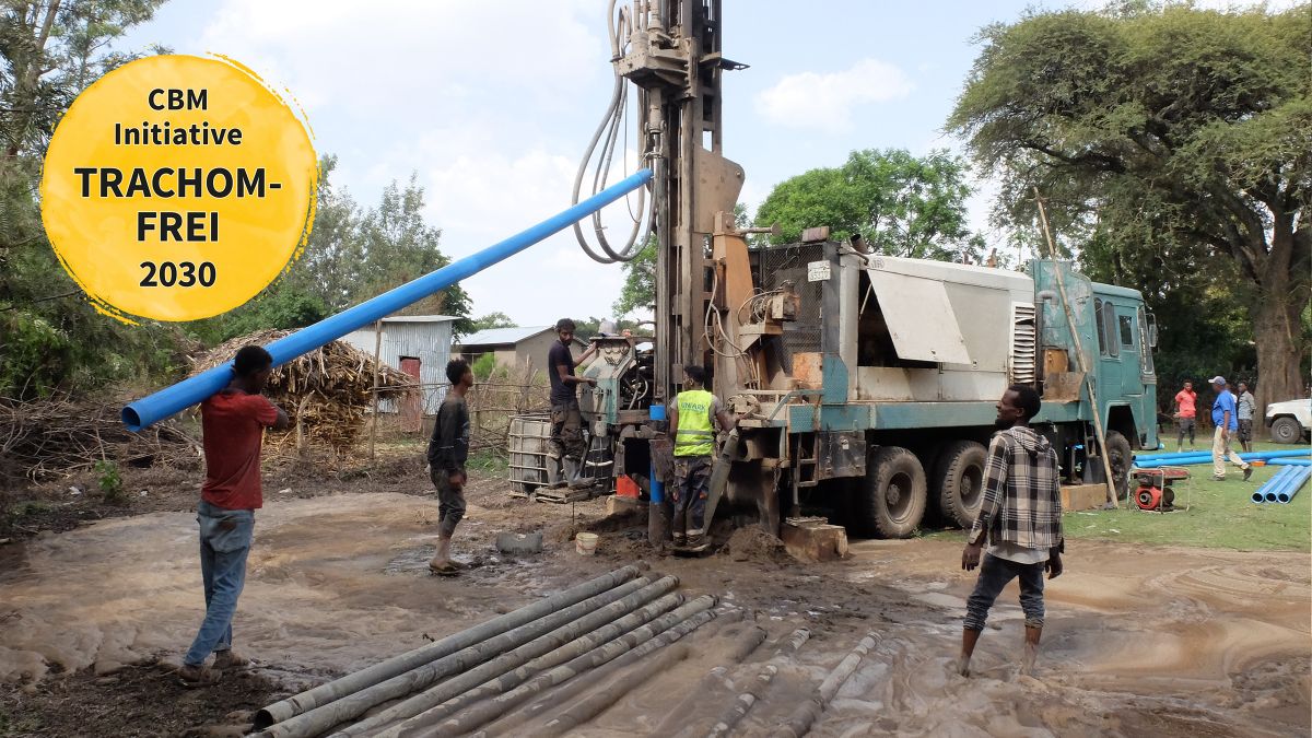 Auf einem Laster steht eine meterhohes Bohrgerät. Männer stehen an dem Gerät, das sich in den Boden schraubt. Auf einem gelben Punkt steht: CBM-Initiative TRACHOMFREI 2030