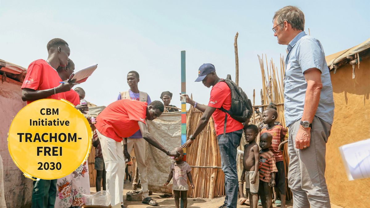 Im Freien zwischen zwei Hütten hält ein Mann eine Messlatte hinter ein Kind, ein anderer gibt dem Kind etwas zu trinken. Weitere Kinder, Männer und Frauen stehen daneben.