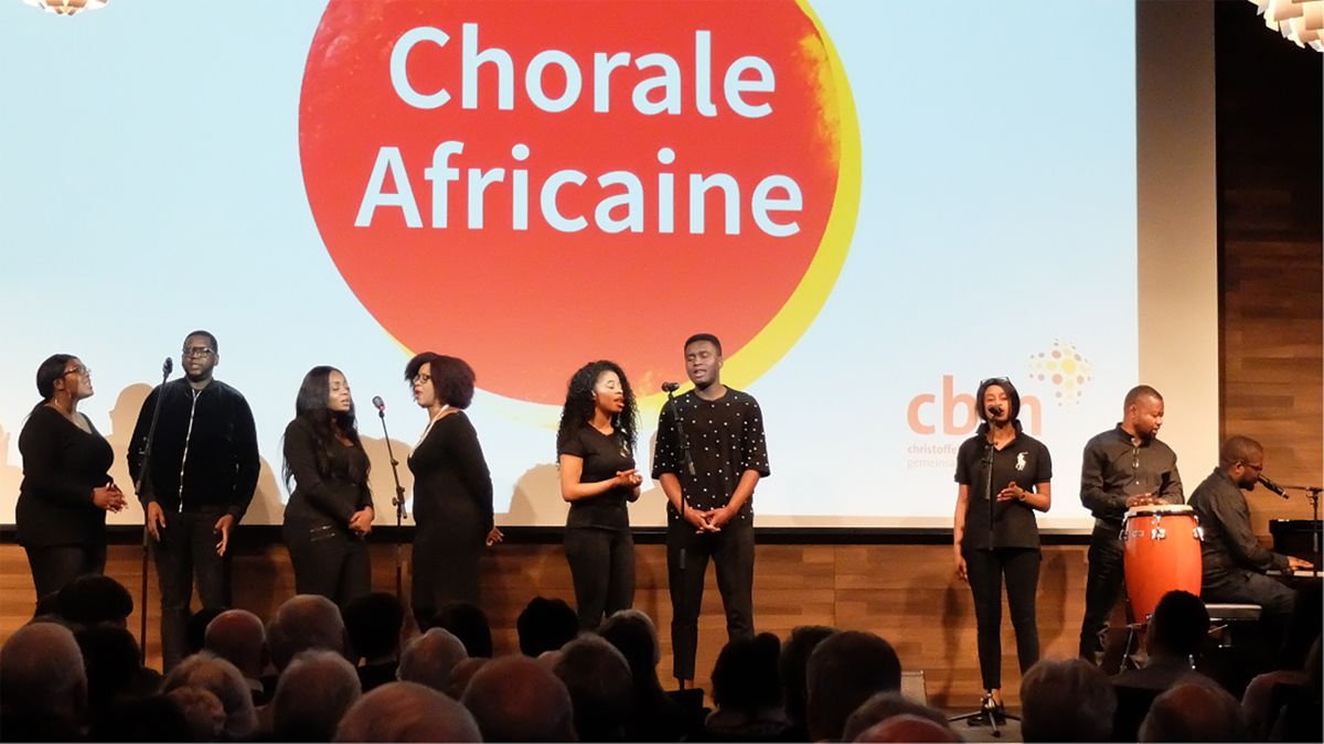 Acht afrikanische Frauen und Männer singen in Mikrofone, hinter ihnen an die Wand projziert ein roter Punkt mit der Aufschrift &quot;Chorale Africaine&quot;
