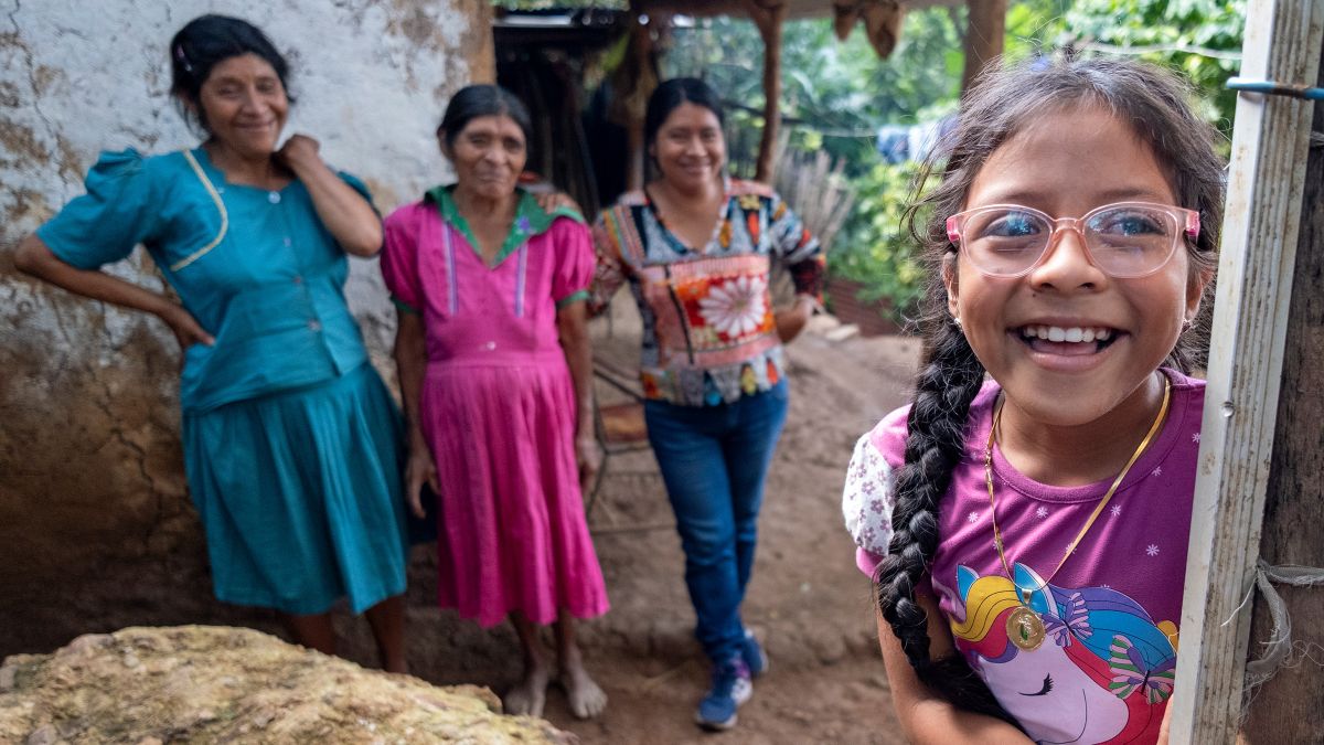 Ein etwa achtjähriges Mädchen mit Zöpfen und Brille steht lachend vor einer Hütte, vor der außerdem drei Frauen stehen.