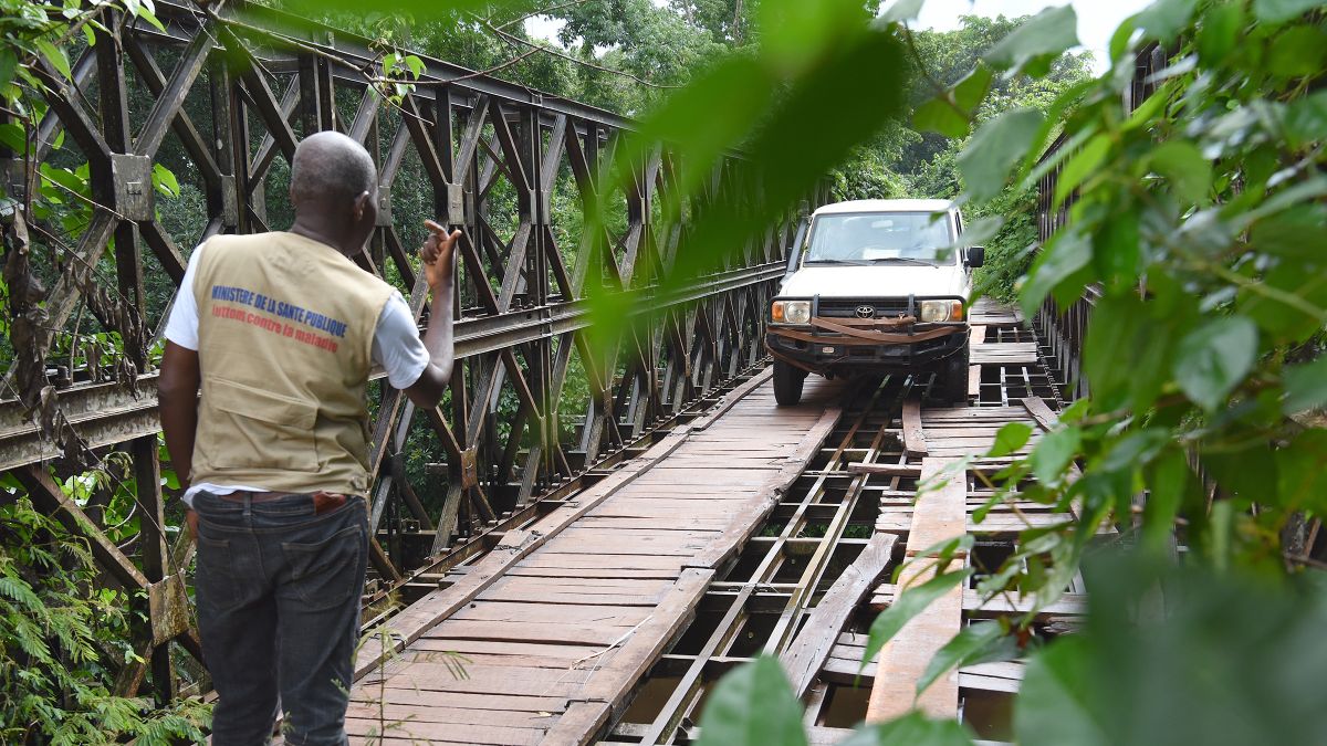 Ein Auto fährt über eine Brücke, deren Boden nur teilweise mit Brettern bedeckt ist und größe Löcher aufweist. Ein Mann steht etwa 20 Meter vorm Auto und gibt dem Fahrer Zeichen, wo er sicher fahren kann.