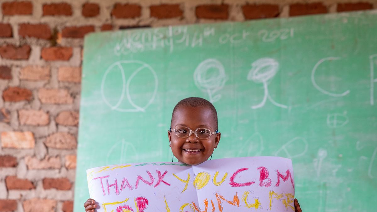 Ein Mädchen sitzt vor einer Tafel und hält ein Bild in der Hand auf dem steht: Thank you CBM