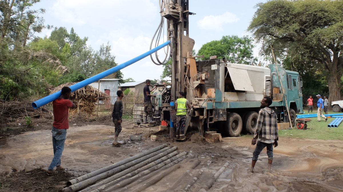 Ein auf einen Lastwagen montierter Bohrer geht in schlammigen Boden, zwei Männer bedienen Maschinen am Bohrer, ein weiterer stemmt ein etwa vier Meter langes Rohr an die Bohrmaschine.