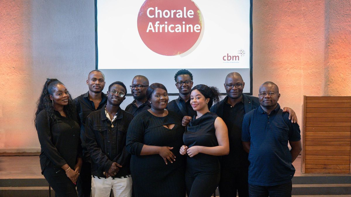 Eine Gruppe aus drei Frauen und sechs Männern in schwarzer Kleidung, an der Wand hinter ihr steht &quot;Chorale Africaine&quot;.