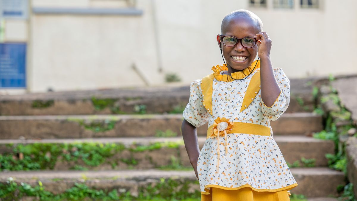 Ein etwa siebenjähriges lachendes Mädchen in gelbgeblümten Rüschenkleidchen steht vor einer Außentreppe. Es trägt eine Brille.