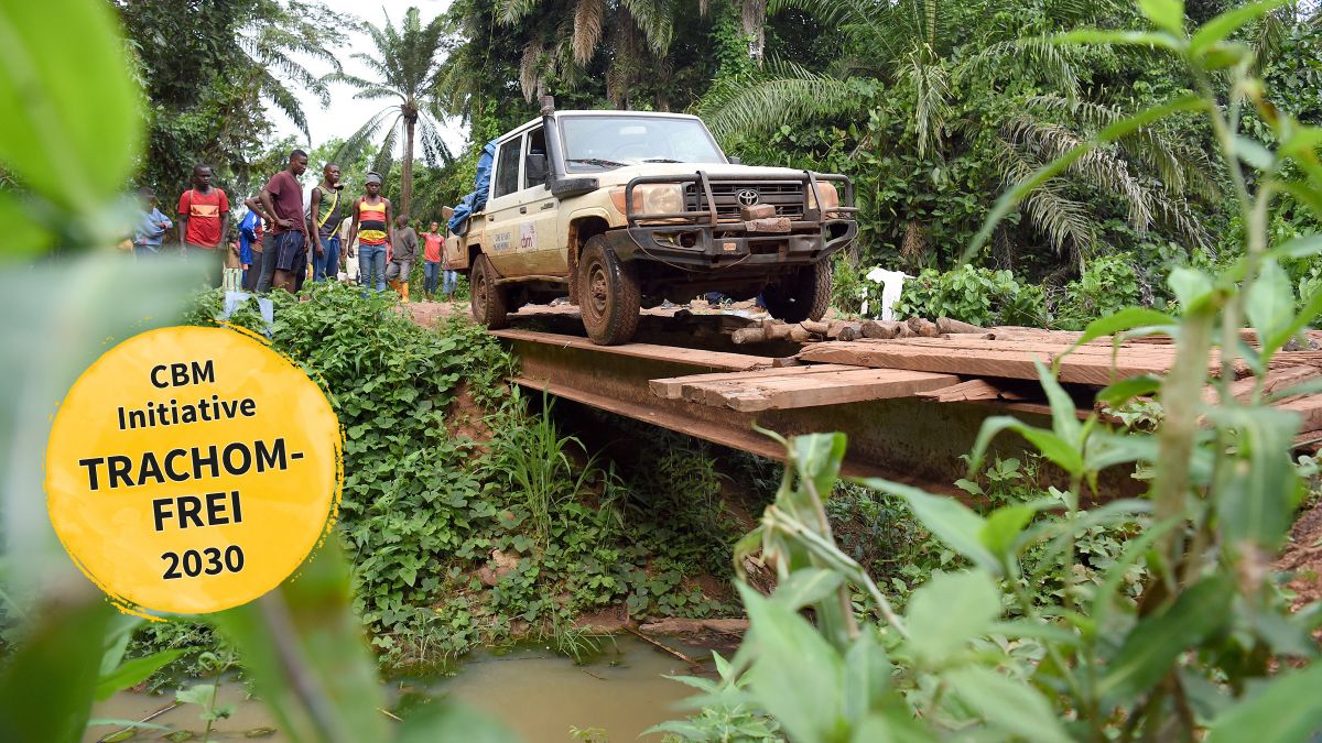 Ein Jepp fährt über eine Brücke, die aus zwei Stahlträgern und ein paar quergelegten, größtenteils fehlenden Brettern besteht. Im Hintergrund stehen einige junge Männer und schauen zu.