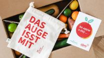 Offener Pappkarton voller Obst und Gemüse, darauf ein Stoffbeutel mit der Aufschrift "Das Auge isst mit"