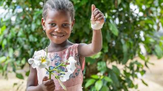 Ein etwa sechjähriges Mädchen hält eine Rispe mit weißen Blüten in der Hand. Die andere Hand hält es hoch, daran ist ein Verband, aus dem das Verschlusselement einer Venenkanüle herausschaut.