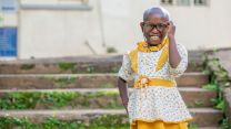 Ein etwa siebenjähriges lachendes Mädchen in gelbgeblümten Rüschenkleidchen steht vor einer Außentreppe. Es trägt eine Brille.