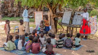 Eine Gruppe von Kindern im Alter zwischen etwa fünf bis sieben Jahren sitzen nebeneinander im Freien auf dem Boden. Eine Frau vor ihnen zeigt auf eine dort aufgestellte Tafel. Ein Mann steht daneben mit Stift und Schreibblock.