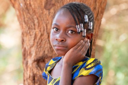 Ein Mädchen mit kleinen Zöpfchen und Glasperlen im Haar steht vor einem Baum. Es legt die Hand ans linke Ohr.
