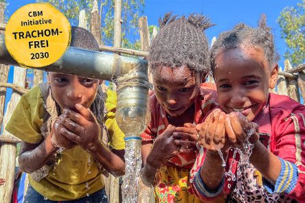 Ein Wasserstrahl schießt aus einem Rohr, daneben waschen sich drei Kinder das Gesicht.