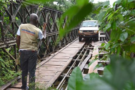 Ein Auto fährt über eine Brücke, deren Boden nur teilweise mit Brettern bedeckt ist und größe Löcher aufweist. Ein Mann steht etwa 20 Meter vorm Auto und gibt dem Fahrer Zeichen, wo er sicher fahren kann.