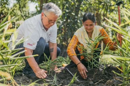 Eine Frau im Sari mit Stock und eine Frau in Jeans und Polohemd hocken auf einem Feld und klopfen mit den Händen die Erde um kleine Grünpflanzen fest.