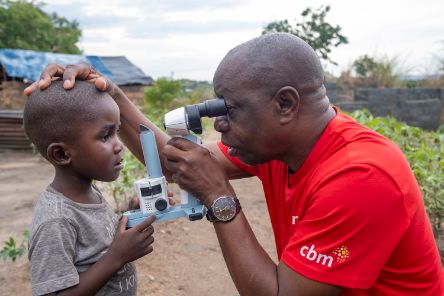 Ein etwa dreijähriger Junge wird von einem Mann im Freien mit einem tragbaren Mikroskop an den Augen untersucht.