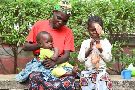 Ein Frau sitzt mit einem Baby auf dem Schoss auf einer Steinbank. Daneben sitzt ein kleines Mädchen mit einem Augenverband. Sie hält eine Tüte in ihren Händen und spielt am Verschluss herum.