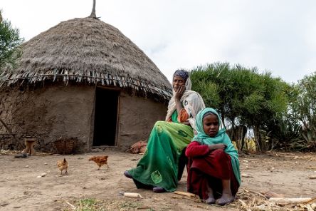 Etwa etwa neunjähriges Mädchen und eine Frau sitzen vor einer Rundhütte. Hühne laufen um sie herum.