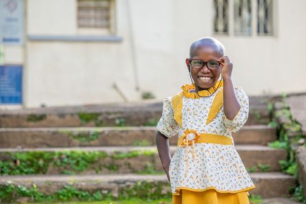 Ein etwa siebenjähriges lachendes Mädchen in gelbgeblümten Rüschenkleidchen steht vor einer Außentreppe. Es trägt eine Brille.
