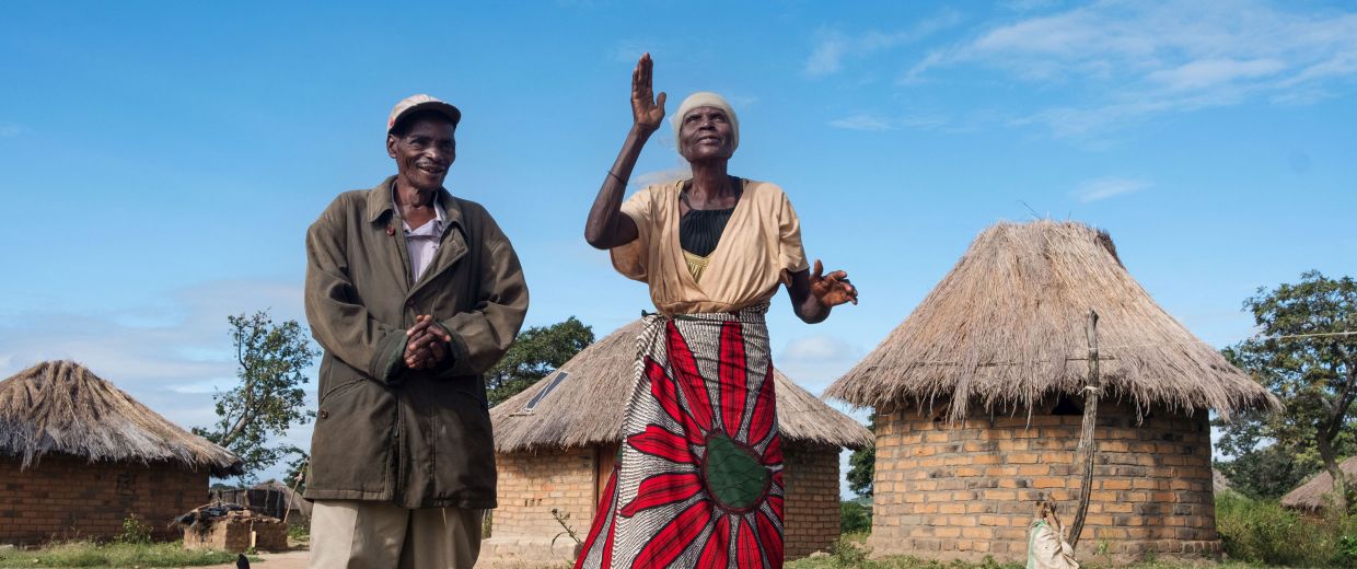 Älteres afrikanisches Paar, lachend und tanzend vor strohgedeckten Rundhütten