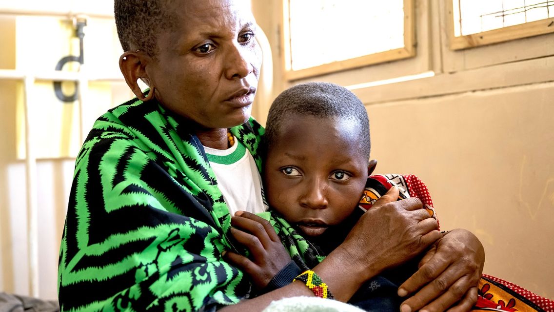 Ein etwa zehnjähriges Mädchen mit hellen Pupillen schmiegt sich an eine Frau in Massai-Tracht.