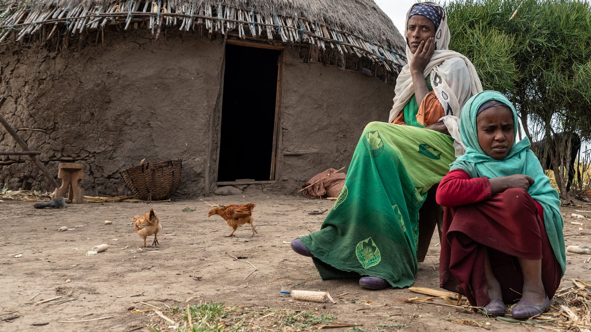 Etwa etwa neunjähriges Mädchen und eine Frau sitzen vor einer Rundhütte. Hühne laufen um sie herum.