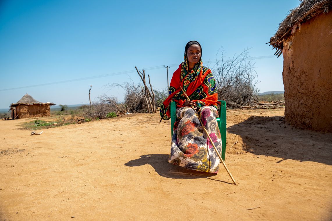 Eine junge Frau sitzt auf einem Plastikstuhl neben einer strohgedeckten Rundhütte.