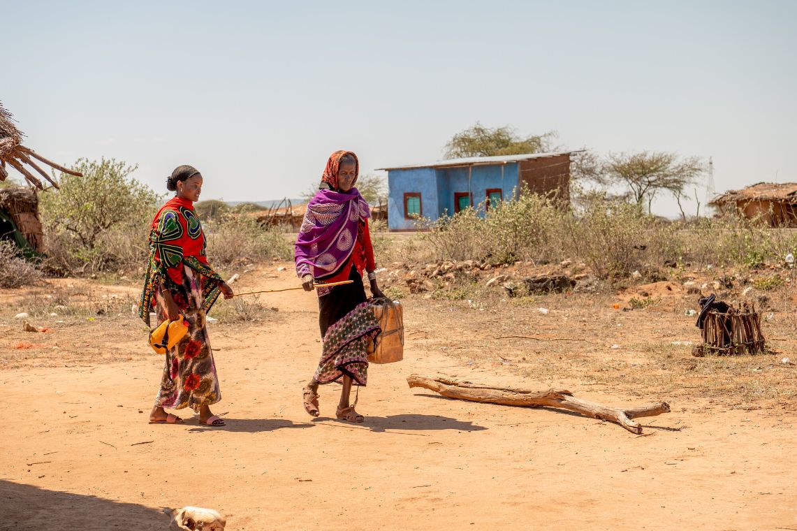 Zwei Frauen, die beide Plastikkanister tragen, gehen hintereinander. Sie halten beide einen Stock zwischen sich an der Hand, der sie verbindet.
