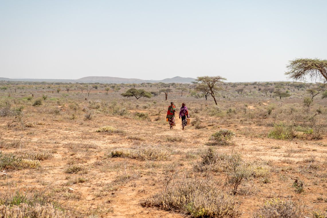 Zwei Frauen in weiter Ferne gehen durch eine trockene Savanne mit spärlicher Vegetation.