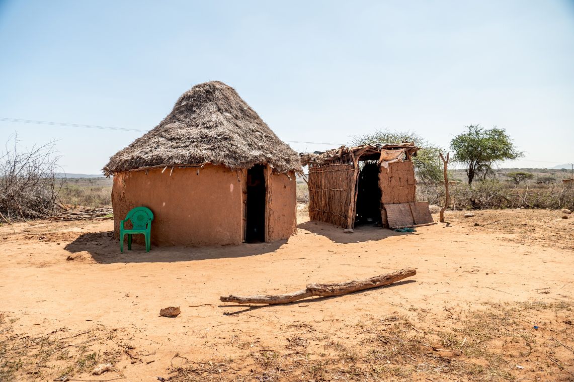 Neben einer Rundhütte steht ein kleiner Stall. Es ist sonnig, der gelbe Boden sieht trocken aus.