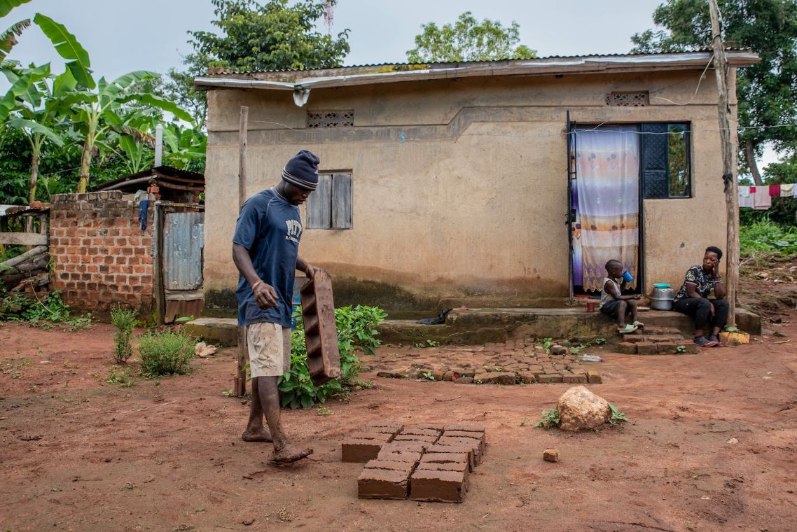 Ein junger Mann in Shorts, T-Shirt und Mütze steht vor einem kleinen Haus mit Wellblechdach im Matsch. Er hält eine Holzform in der Hand, vor ihm auf dem Boden liegen frisch geformte Backsteine.