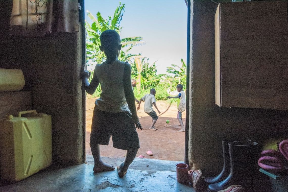 Ein etwa vierjähriger Junge mit etxremen O-Beinen steht im Eingang der Hütte und schaut nach draußen, wo andere Kinder Fußball spielen.