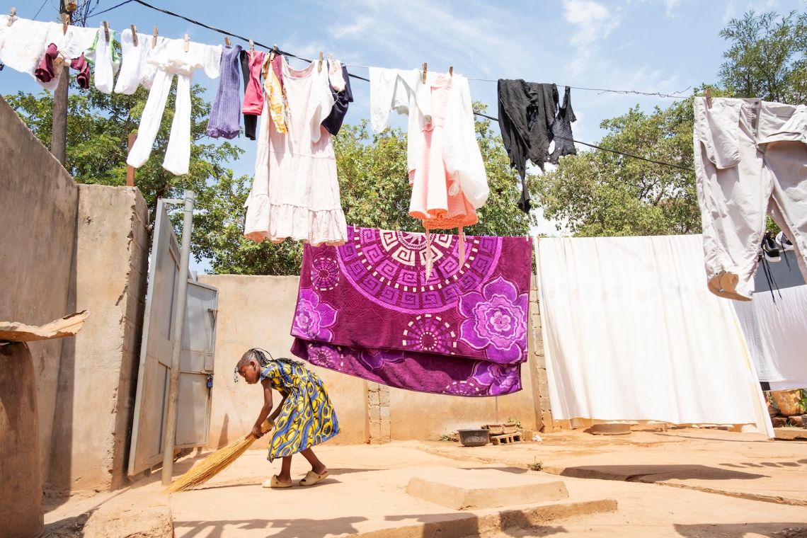 Ein kleines Mädchen fegt mit einem Reisigbesen einen Hof, vor dem eine Leine gespannt ist, an der Wäsche flattert.