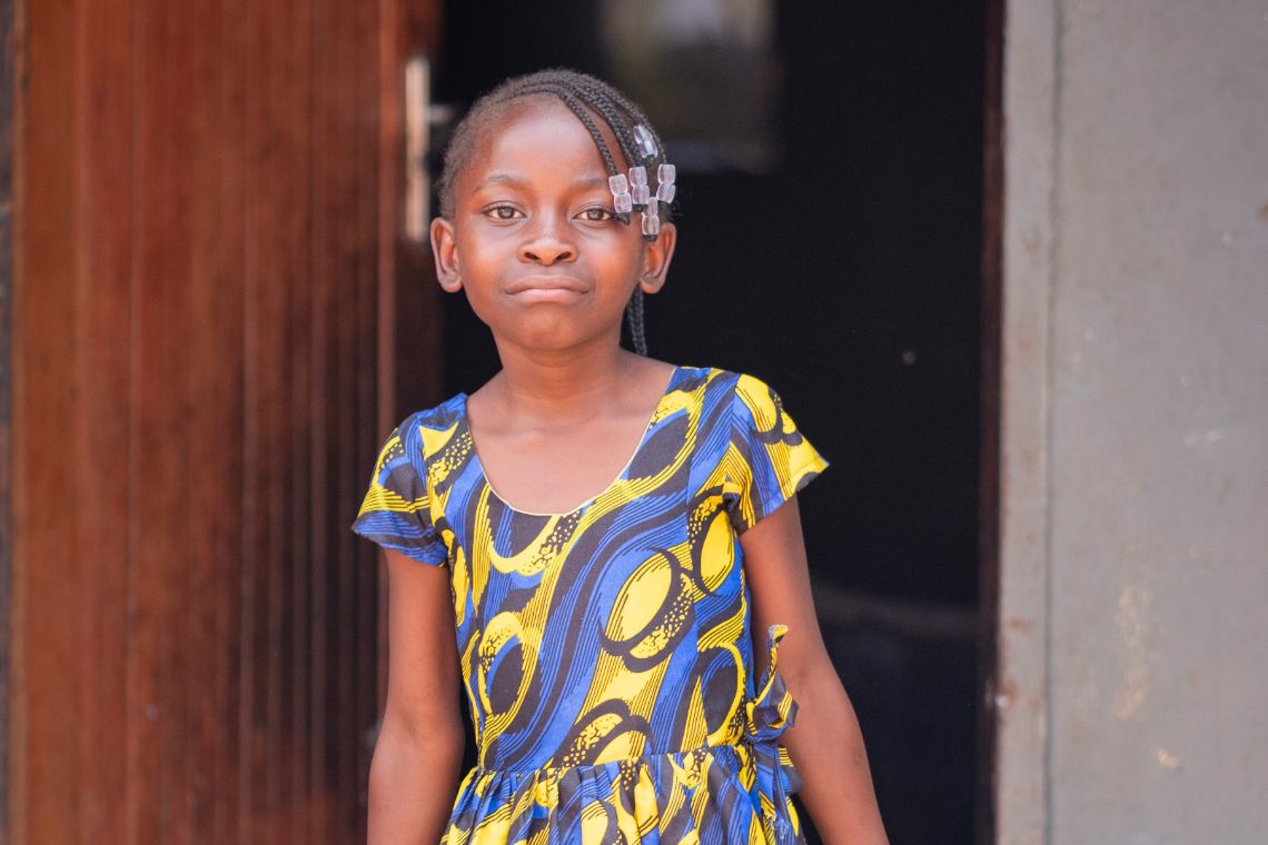 Ein kleines Mädchen fegt mit einem Reisigbesen einen Hof, vor dem eine Leine gespannt ist, an der Wäsche flattert.