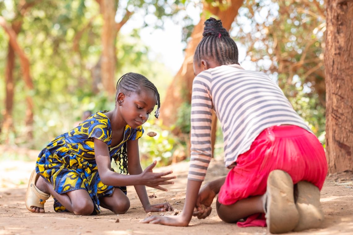 Ein etwa Neunjährige und etwa Zwölfjährige knieen am Boden und spielen mit Steinchen.