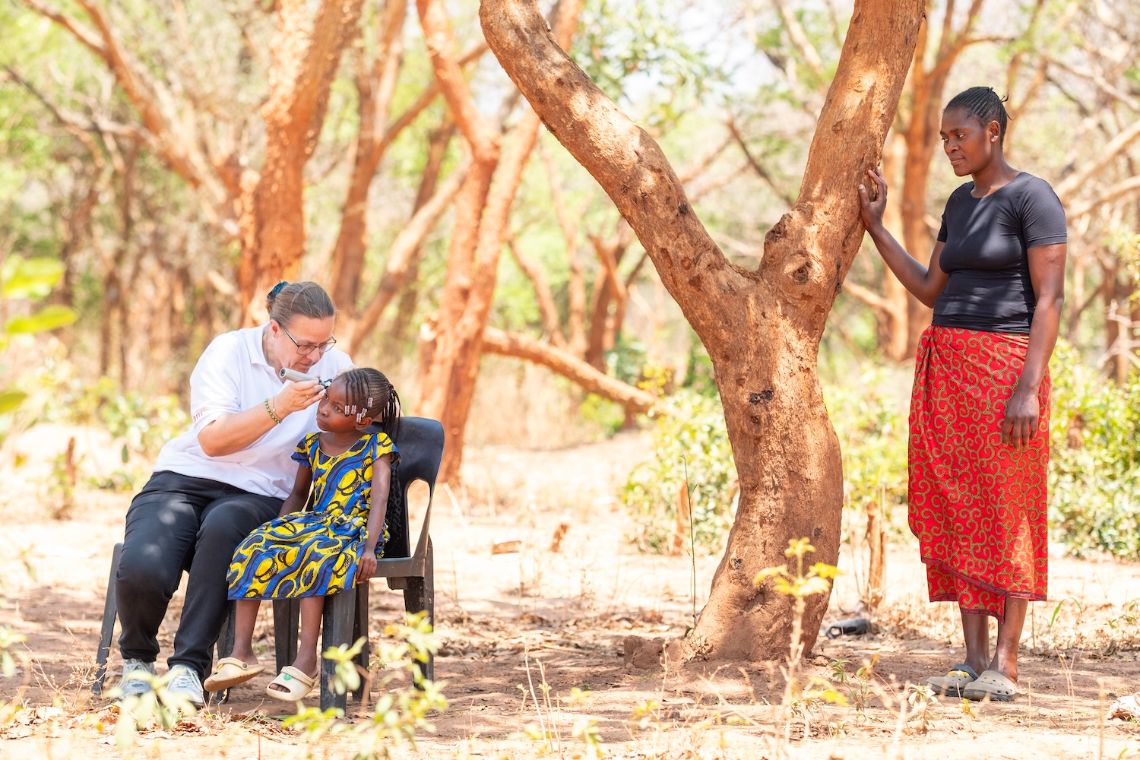 Eine Frau im weißen Polohemd und ein etwa neunjähriges Mädchen sitzen unter Bäumen auf Plastikstühlen nebeneinander. Die Frau untersucht das Ohr des Mädchens. Eine weitere Frau mit buntem Rock steht wenige Schritte daneben und schaut zu.