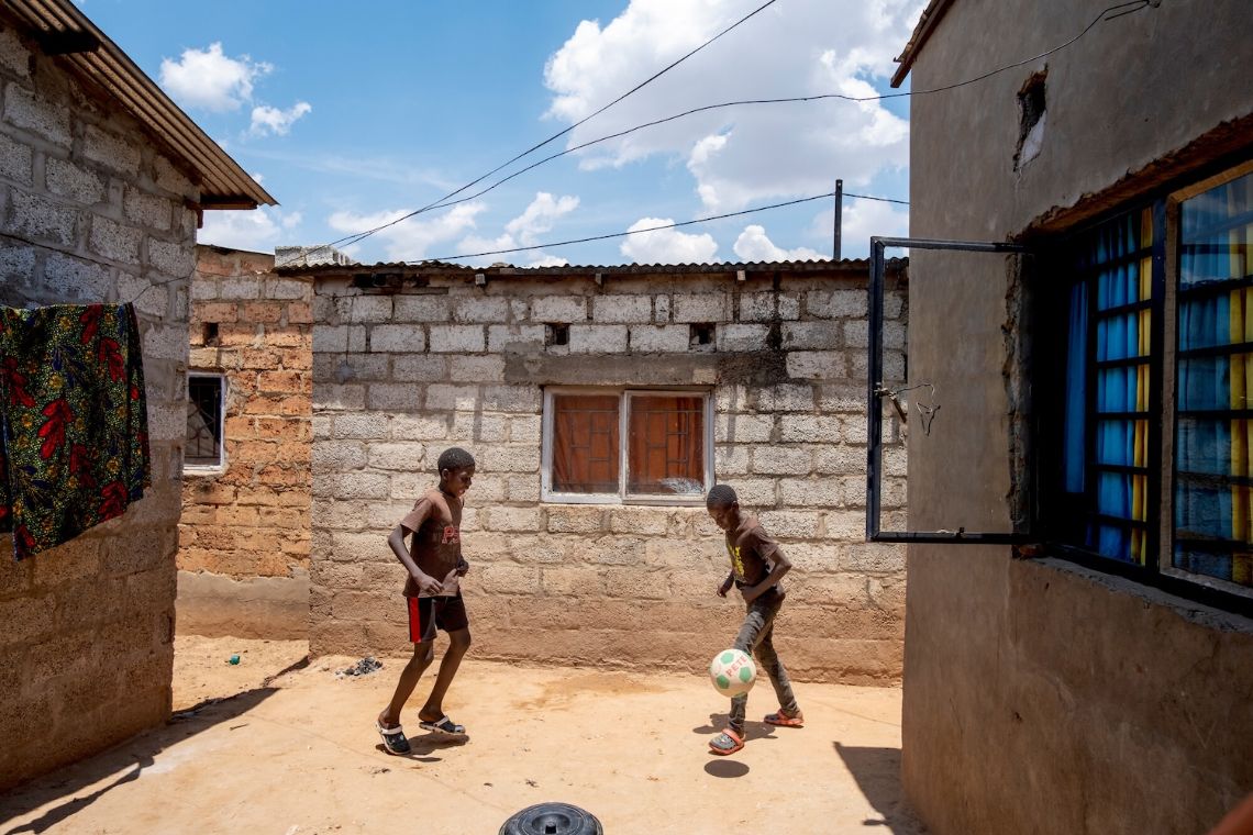 Zwei etwa zwölfjährige Jungen spielen zwischen eingeschossigen, unverputzen kleinen Häusern Fußball.