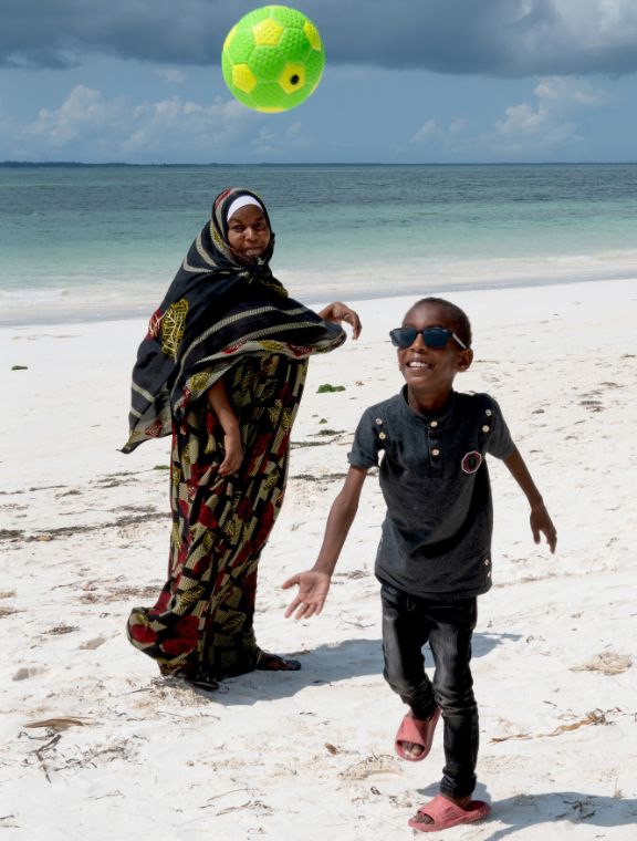 Ein lachender, etwa achtjähriger Junge mit Brille und eine Frau mit langem Gewand und Kopftuch an einem Sandstrand: Der Junge schaut einem Fußball nach, der fast über ihm fliegt.