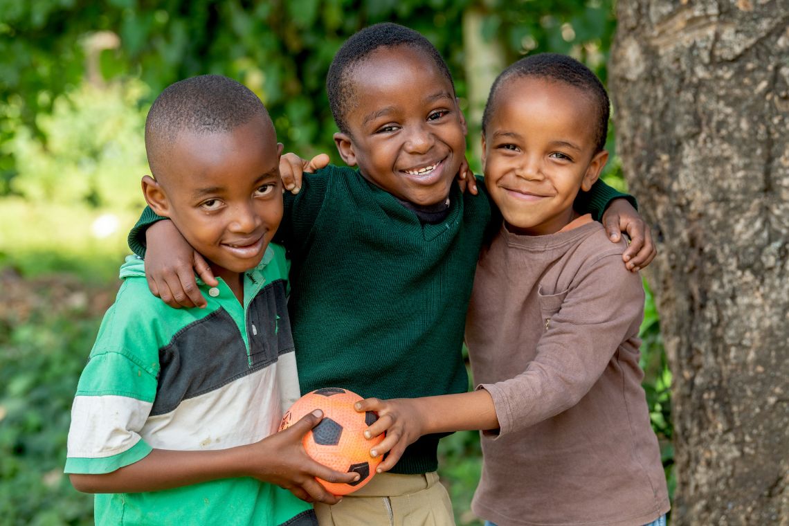 Ein etwa achtjähriger Junge legt die Arme um die Schultern der Buben rechts und link von ihm, die zusammen einen Ball festhalten.