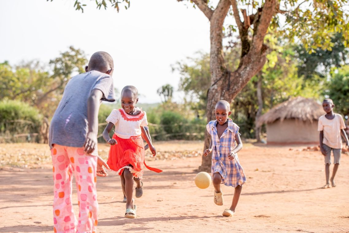 Zwei kleine Mädchen laufen einem Ball nach, zwei weitere Kinder stehen dabei.