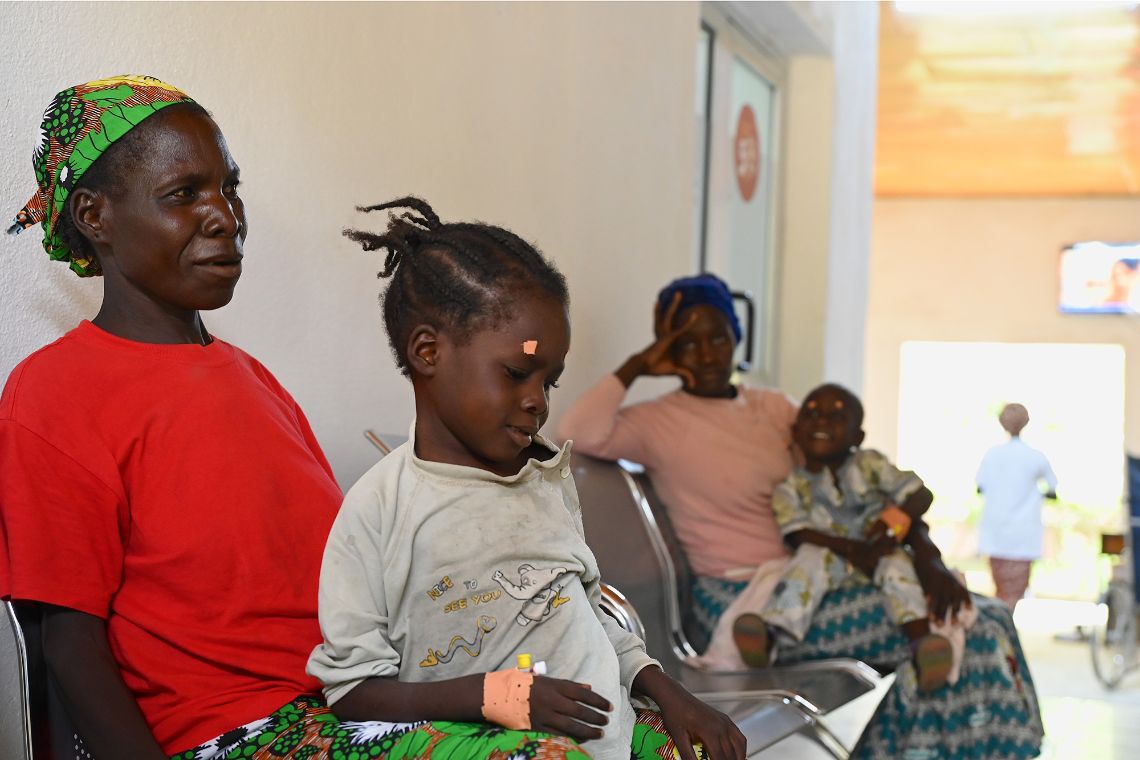 Eine Frau mit buntem Kopftuch hat ein etwa achtjähriges Mädchen auf dem Schoß, über dessen Auge ein kleines Pflaster auf der Stirn klebt.