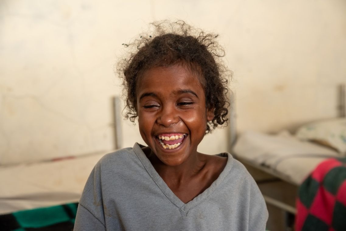 Etwa neujähriges, lachendes Mädchen, an dessen Wange noch ein herunterhängender Verband klebt.